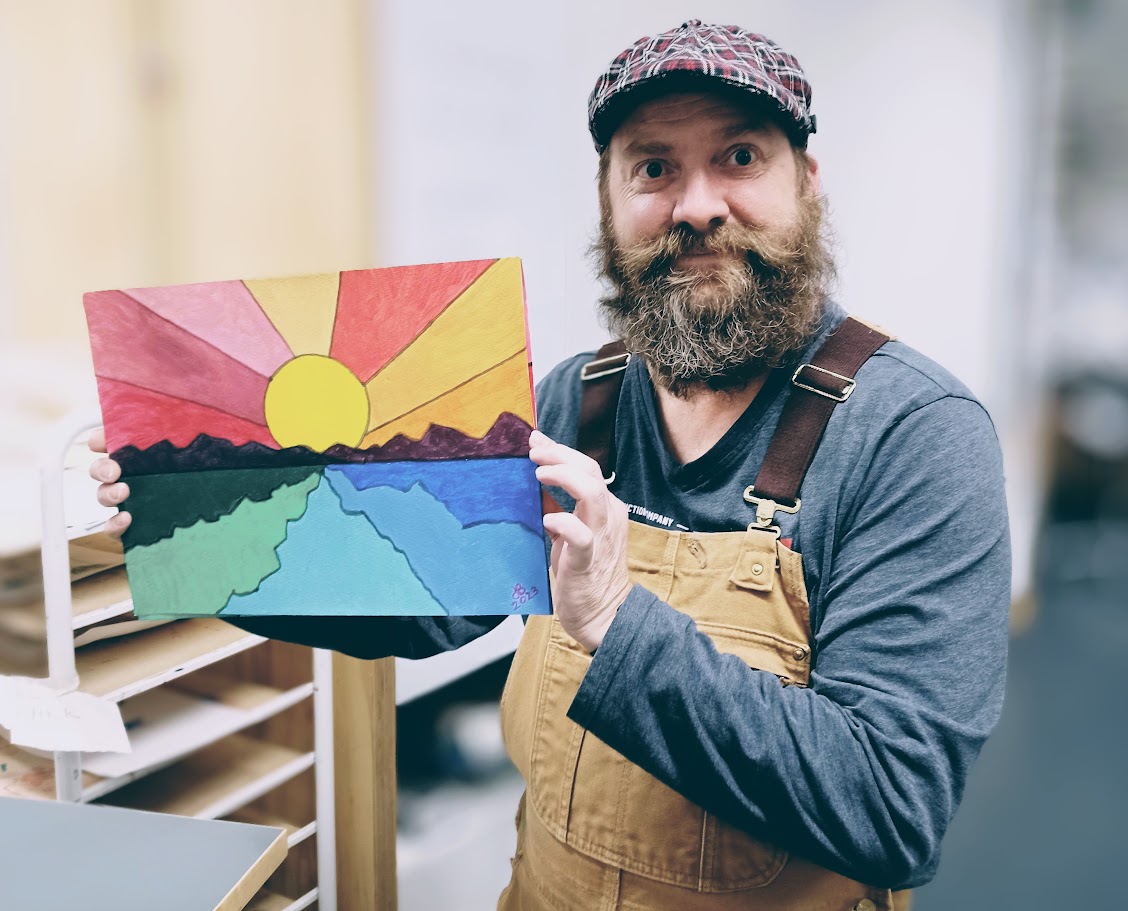 kuvassa Justin Ballew, joka on parrakasmies, hattu päässä ja oppilaan maalaama taideteos kädessä