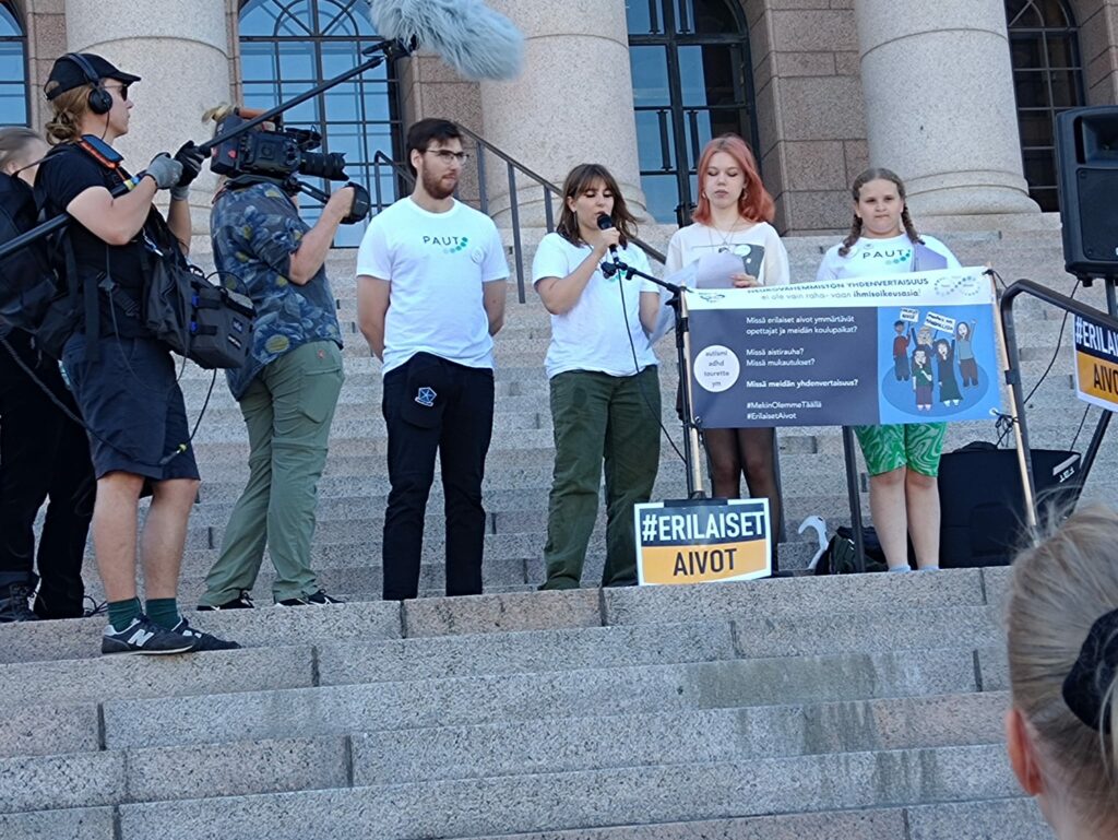 Nuoret aktivistit ryhmän jäseniä seisoo mikrofonin edessä eduskuntatalon portaille. Heidän ympärillään on kuvausryhmä.